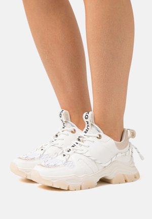 Baskets blanches épaisses avec semelles beige épaisses et étiquettes de languette de marque, portées pieds nus sur un fond clair uni.