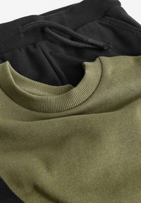 Olivengrøn sweatshirt med ribbet krave, parret med sorte sweatpants med elastisk talje og snor. Glat tekstur.