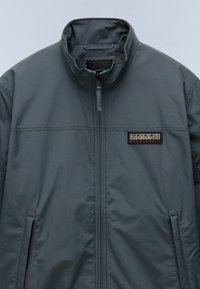 Giacca grigia con colletto alto, dal tessuto liscio, chiusura con zip e tasche laterali. Presenta un logo del marchio ricamato sul petto.