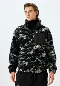 Zwart-wit fleece pullover met een ritszak, hoge kraag en een gestructureerd patroon. Voorzien van elastische boorden en een trekkoord aan de onderkant.
