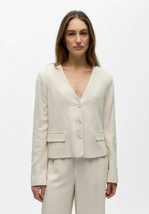 Femme portant un blazer boutonné couleur crème avec poches à rabat et un pantalon assorti, debout devant un fond blanc uni.