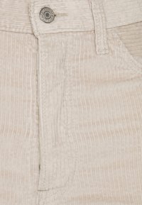 Lichtbeige corduroy stof met verticale strepen, voorzien van een zilveren knoopsluiting en gestikte zakken op de voorkant.