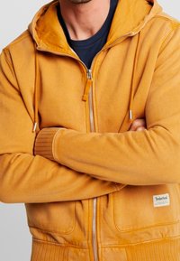 Sudadera con cremallera en color naranja, con puños y dobladillo acanalados. Incluye cremallera plateada y cordones. La tela parece suave con un acabado texturizado. Etiqueta de Timberland visible.