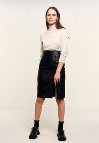 Zwarte leren kokerrok met een voorste split, gecombineerd met een beige coltrui. Model draagt zwarte schoenen met dikke zolen.