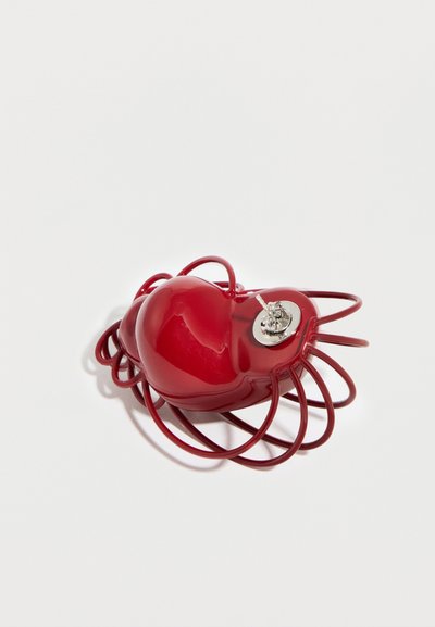 Sijajni rdeč predmet v obliki srca s zankastim žičnim okvirjem in majhnim kovinskim stikalom na eni strani, postavljen na belo površino.