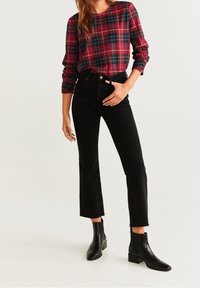 Vrouw in een rood en marineblauw geruite blouse met lange mouwen, hoge zwarte cropped jeans en zwarte enkellaarzen, staande tegen een eenvoudige achtergrond.