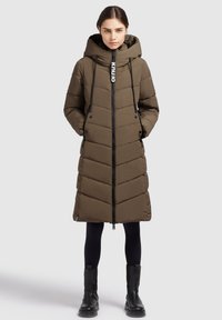 Naine, kes kannab pikka oliiviroheline puffer-katet, millel on kapuuts, musti legginsid ja musti saapaid, seisab hallil taustal, käed taskutes.