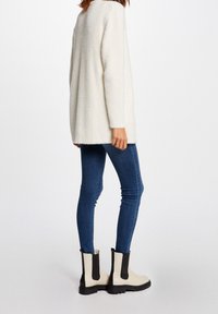 Vrouw draagt een wit zacht langärmelig truitje, blauwe strakke spijkerbroek en witte enkellaarzen met zwarte elastische zijkanten.