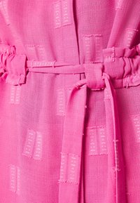 Tessuto rosa con una trama testurizzata e motivi rettangolari. Una cintura in tessuto coordinato è annodata in vita, evidenziando la struttura del capo.