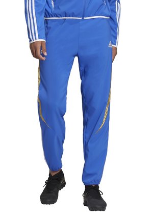 Blauwe Adidas trainingsbroek met gouden zijstrepen, elastische boorden en zwarte sportschoenen, gedragen met een bijpassende blauwe jas.