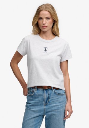 Vrouw met blond haar die een wit T-shirt met ankerlogo en blauwe spijkerbroek draagt, staat met één hand op haar heup.