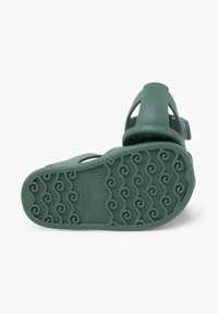 Sándalo de goma verde con suela texturizada que presenta patrones de olas, correa ajustable y parte superior lisa con agujeros de ventilación.