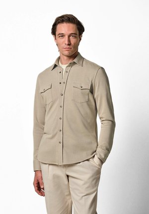 Mann steht mit der Hand in der Tasche, trägt ein beiges Hemd mit Brusttaschen und helle Hosen vor einfarbigem Hintergrund.