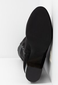 Bottine en daim noire avec un petit talon carré, semelle tournée vers l'avant, inclinée contre un fond blanc en angle.