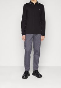 Long-sleeve black polo shirt with white collar accents, paired with gray pants and black shoes, standing against a neutral background.