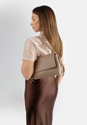 Donna con capelli castano lisci, indossa una blusa color crema e una gonna marrone, con una borsa a tracolla in pelle color taupe sulla spalla destra.