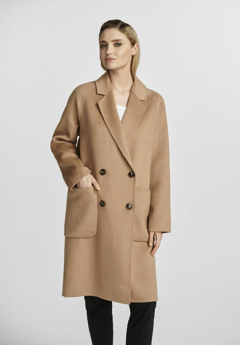 Płaszcz z mieszanki wełny w kolorze camel z podwójnym zapięciem, dużymi przednimi kieszeniami i brązowymi guzikami. Posiada klapę w kształcie litery V oraz luźny krój.