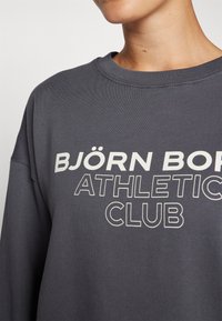Sudadera gris con un cuello redondeado, que presenta un gran texto blanco que dice "BJÖRN BORG ATHLETIC CLUB" en una fuente moderna y en negrita.