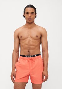 Korallfargede svømmetights med elastisk midje og snøring, med en svart Calvin Klein-logo stripe i midjen. Glatt, lett stoff.