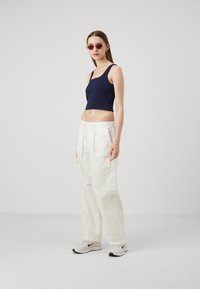 Top court en marine avec des fines bretelles, associé à un pantalon cargo blanc ample avec des poches latérales. Le modèle porte des lunettes de soleil roses.