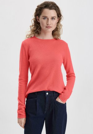Korallefarbener Strickpullover mit langen Ärmeln, runder Ausschnitt und weicher Textur. Kombiniert mit dunkelblauen Jeans, die Taschen und einen Knopf haben.