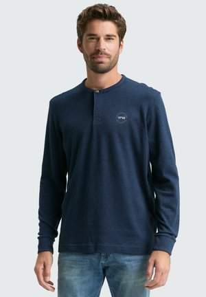 Man in een marineblauwe langwerpige henley-shirt met een kleine grafische afbeelding op de linkerborst en lichtblauwe jeans voor een effen achtergrond.