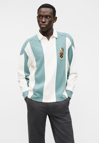 Ριγέ πόλο πουκάμισο σε ανοιχτό μπλε και λευκό, με γιακά, μακριά μανίκια και κεντητό έμβλημα στο στήθος.