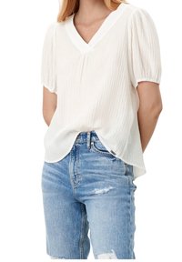 White short-sleeve blouse with a V-neck, textured fabric, and slightly curved hem. Paired with light blue distressed denim jeans.