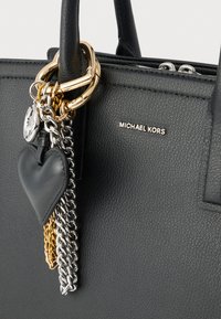 Μαύρη τσάντα χειρός από δερμάτινη υφή με χρυσά και ασημένια μεταλλικά στοιχεία, διακοσμημένη με γοητεία σε σχήμα καρδιάς και αλυσίδες. Παρουσία του λογοτύπου Michael Kors.