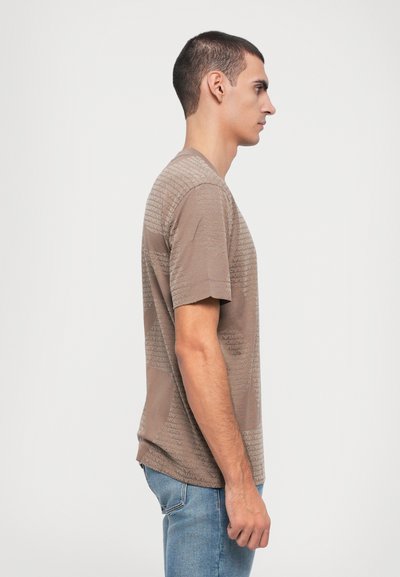 Lys brun kortermet t-skjorte med et teksturert mønster av gjentatte merkenavn, rund hals-design og myk stoff, parret med blå jeans.