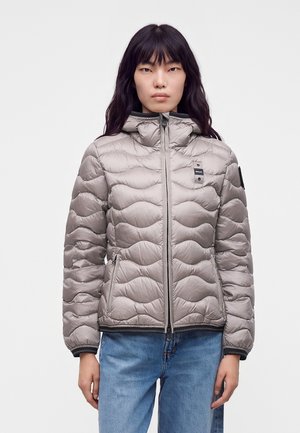 Femme portant une doudoune matelassée gris clair avec capuche et un jean bleu, debout devant un fond blanc uni.