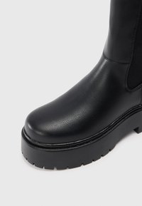 Botă ankle neagră, cu un superior din piele texturată, vârf rotund și talpă groasă din cauciuc negru, cu un model de profil.