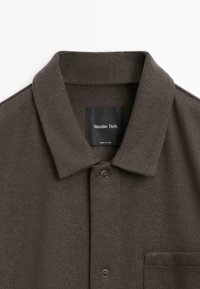 Bruine gebreide polo met een klassieke kraag, een voorzak en een knoopsluiting met zilverkleurige hardware. Merkenlabel zichtbaar aan de binnenkant van de kraag.