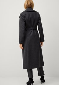 Cappotto lungo in lana grigia scura con un ampio colletto, vita cintrata e vestibilità su misura. Presenta maniche lunghe e una texture liscia con dettagli minimi.