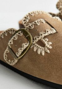 Brown suede sandal featuring a decorative stitched strap with cream-colored threading and a metallic buckle accent on the side.