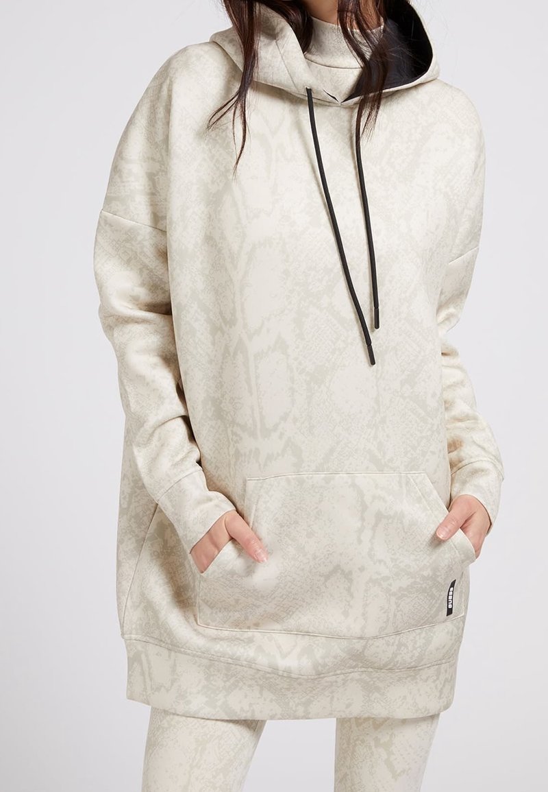 Femme portant un sweat à capuche oversize beige à motif serpent, avec cordons noirs et poche avant, mains partiellement dans les poches.