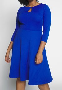 Robe bleu roi avec un col rond et un détail en forme de trou de serrure. Elle présente des manches trois-quarts et une jupe évasée, confectionnée dans un tissu texturé.