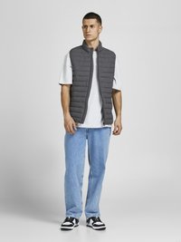 Grau gepolsterte Weste mit hohem Kragen über einem grauen T-Shirt, kombiniert mit hellblauen Jeans und schwarz-weißen Sneakers, stehend vor einem einfarbigen Hintergrund.