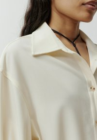 Crème-kleurige blouse met een kraag, knoopsluiting en een gladde, zachte stofstructuur. Bevat een zwart koordaccessoire.