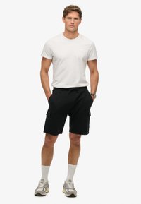 Czarne szorty cargo z przednimi kieszeniami, zestawione z dopasowaną białą koszulką i szarymi trampkami. Model stoi z rękami w kieszeniach.