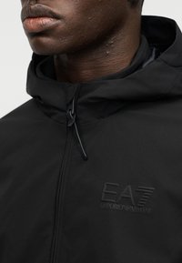 Μαύρο μπουφάν με κουκούλα και υφή, με διακριτικό ανάγλυφο λογότυπο EA7 στο στήθος και φερμουάρ μπροστά.