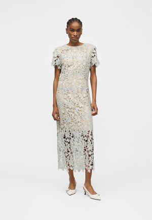 Femme debout portant une robe midi en dentelle florale blanche avec manches courtes et des talons blancs pointus avec petits nœuds.