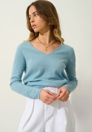 Un pull V en bleu clair, fabriqué dans un matériau doux, associé à un pantalon blanc taille haute. Le pull présente des manches longues et une coupe décontractée.