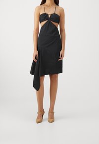 Černé šaty s mašlí a výřezy, s volným asymetrickým lemem a jednoduchými ramínky. Hladká textura a minimalistický design.