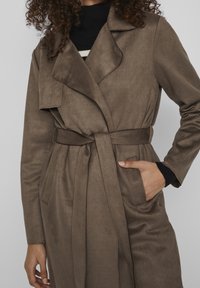 Manteau en daim marron avec ceinture à la taille, col à revers et poches latérales. Présente une texture douce et une finition lisse, idéal pour superposer.