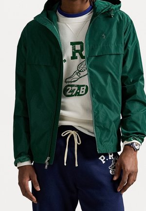Man wearing a dark green hooded zip-up jacket over a white graphic sweatshirt and navy blue sweatpants with white drawstring.