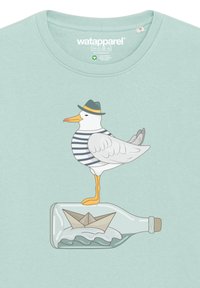 T-shirt vert menthe avec un dessin de mouette cartoon portant une chemise rayée et un chapeau, se tenant sur une bouteille avec un bateau en papier à l'intérieur.