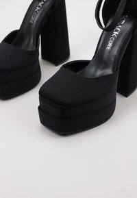 Zapatos de tacón negro con una parte superior de tela suave, tacones de bloque gruesos y una suela delantera ancha, que ofrecen una silueta moderna y estructurada.