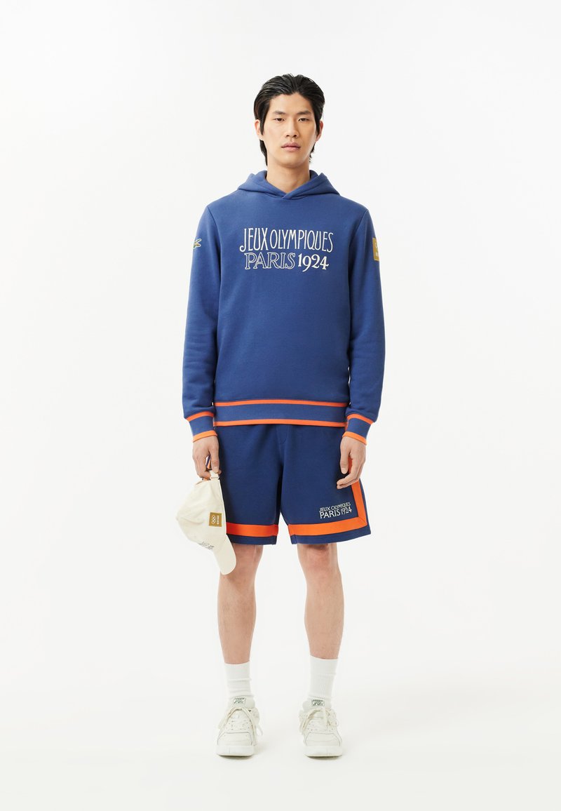 Mann in blauer Kapuzenjacke und Shorts mit dem Text "Jeux Olympiques Paris 1924", der eine beige Kappe hält und vor einem weißen Hintergrund steht.
