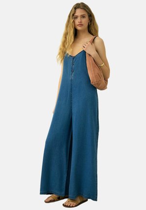 Frau mit langen blonden Haaren, die einen blauen ärmellosen Jumpsuit mit weitem Bein trägt, braune Sandalen anhat und eine hellbraune Umhängetasche trägt.